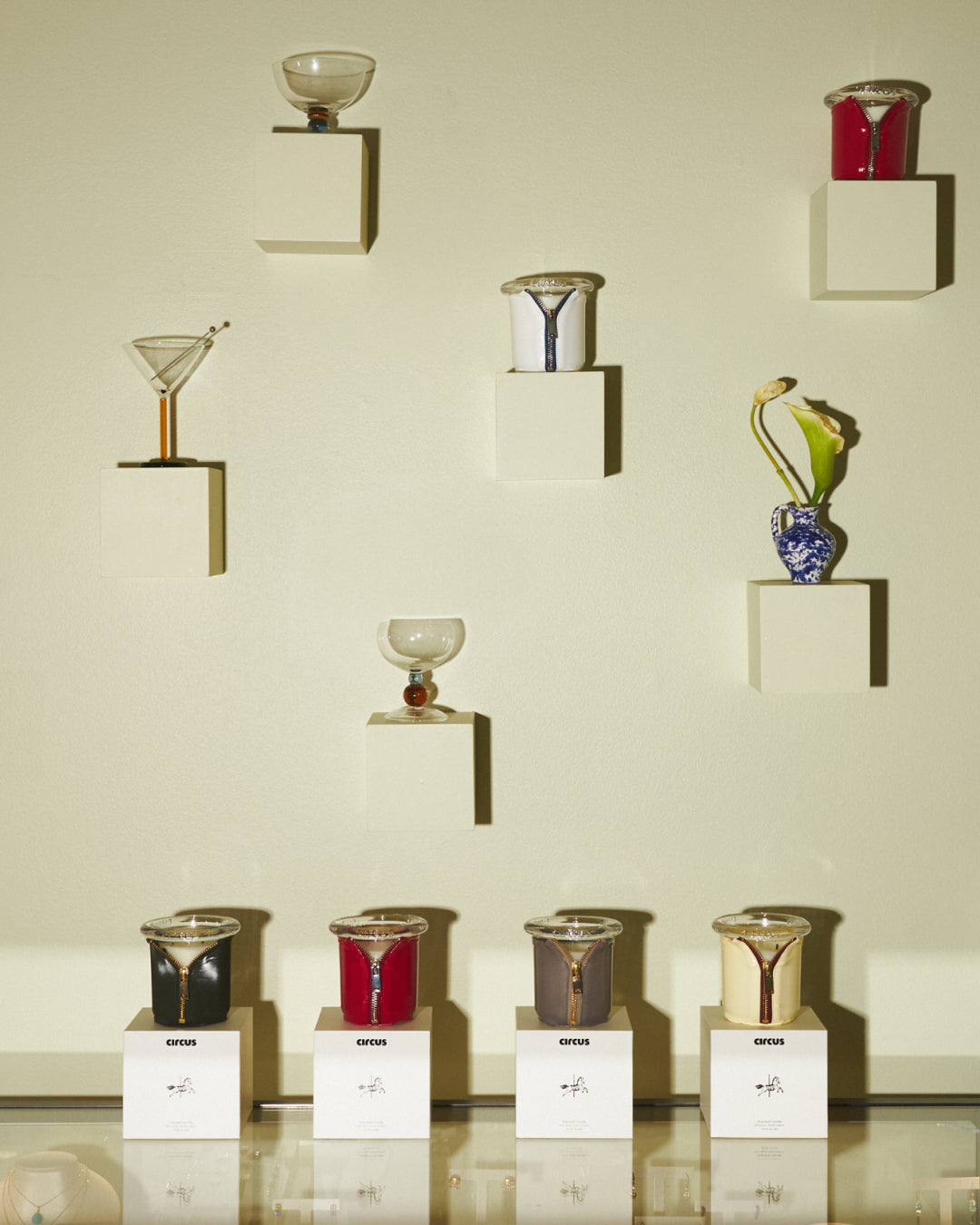 A collection of hand-blown glass vessels, including tumblers, a martini glass, and a vase, displayed on white wall-mounted blocks and a glass shelf against a warm beige background featuring circus candles