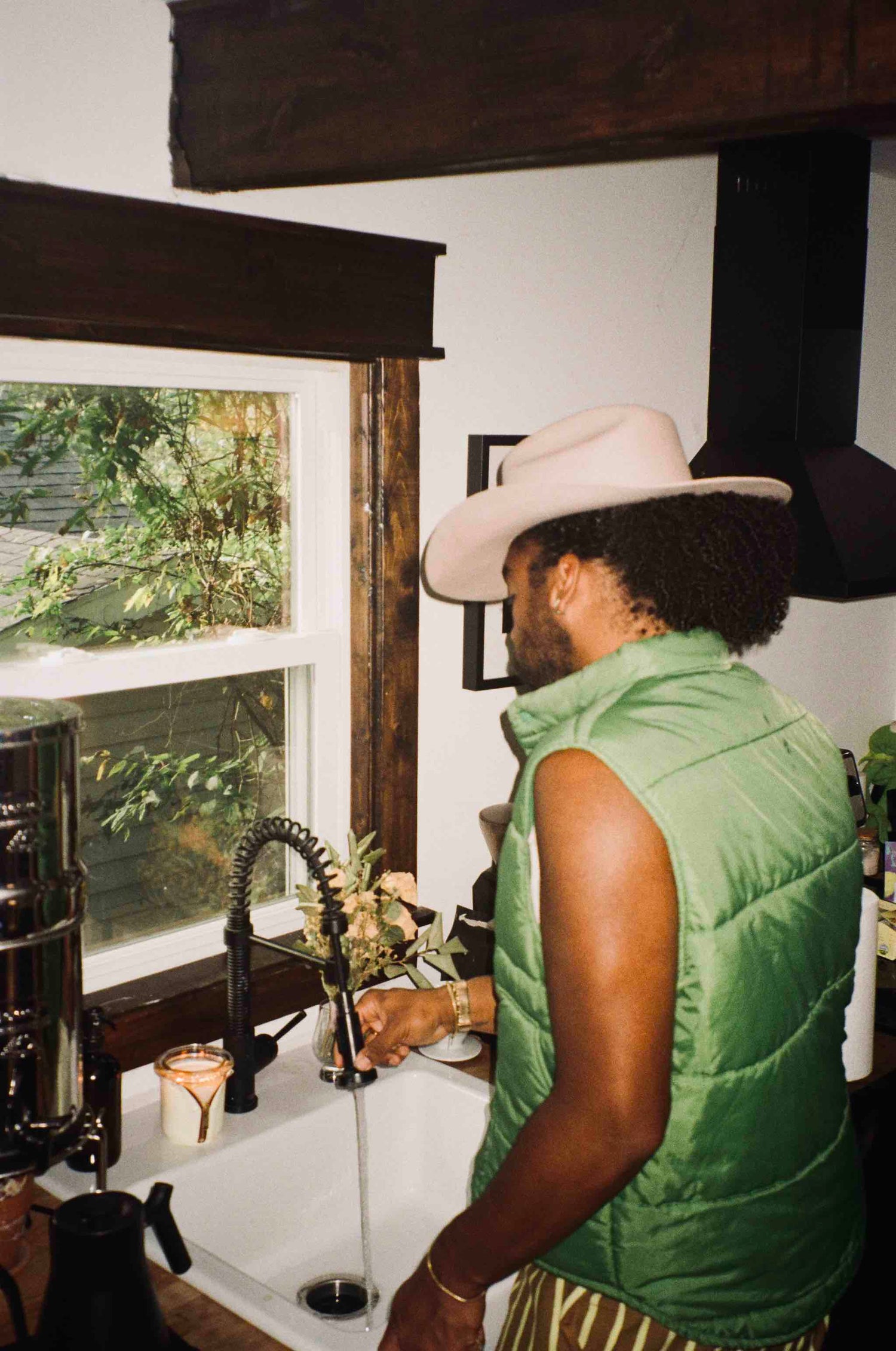 photographer joseph ross wearing a green vest and cowboy hat standing in a kitchen with circus candle bound lit
