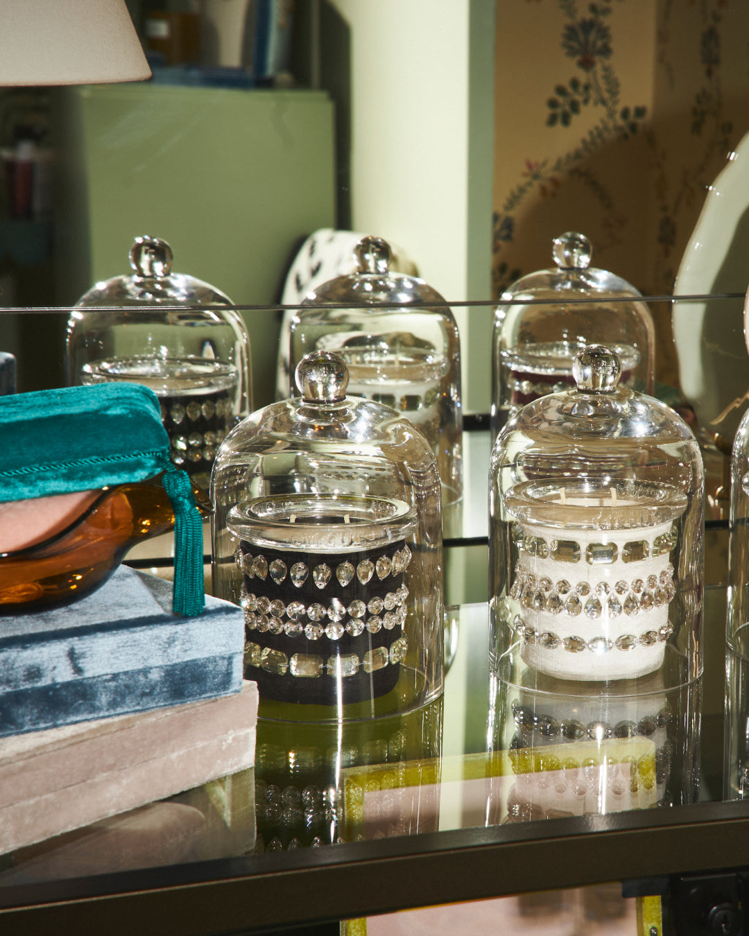 A close-up of several clear glass cloche jars with silver-toned lids, displayed on a reflective glass shelf. Some jars contain Crystal Fetish by Circus candles in black and in white