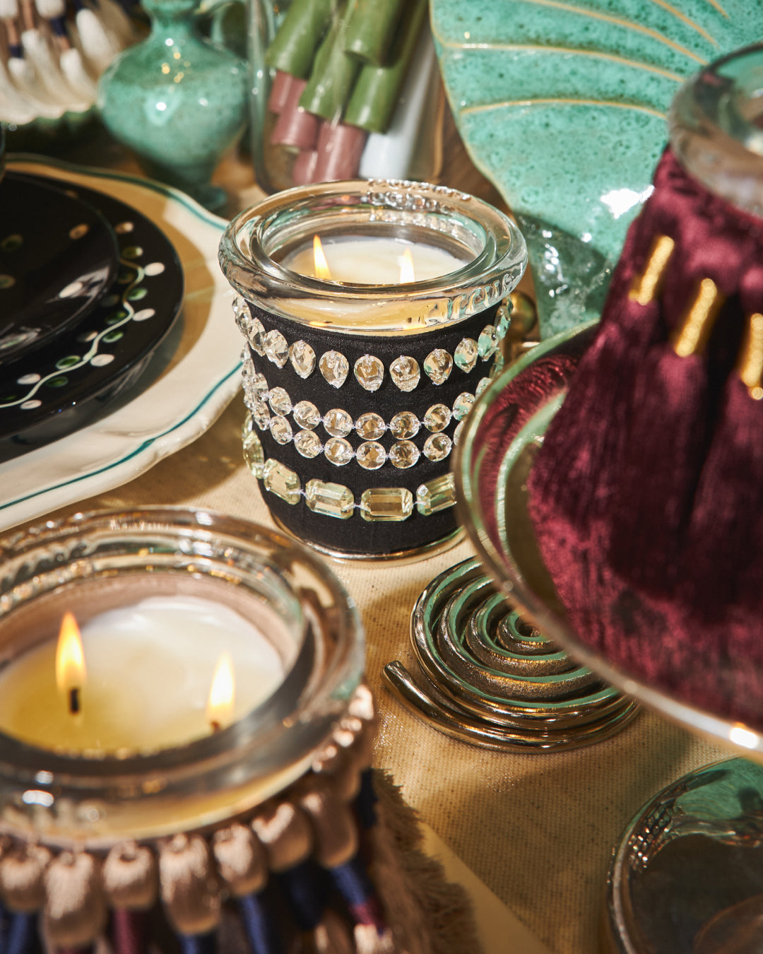 A close-up styled tabletop scene featuring two lit crystal fetish circus candles in decorative holders. The central candle is in a clear glass jar wrapped with black fabric and rows of rhinestones, while the foreground candle has a metallic gold-toned fringed rim.