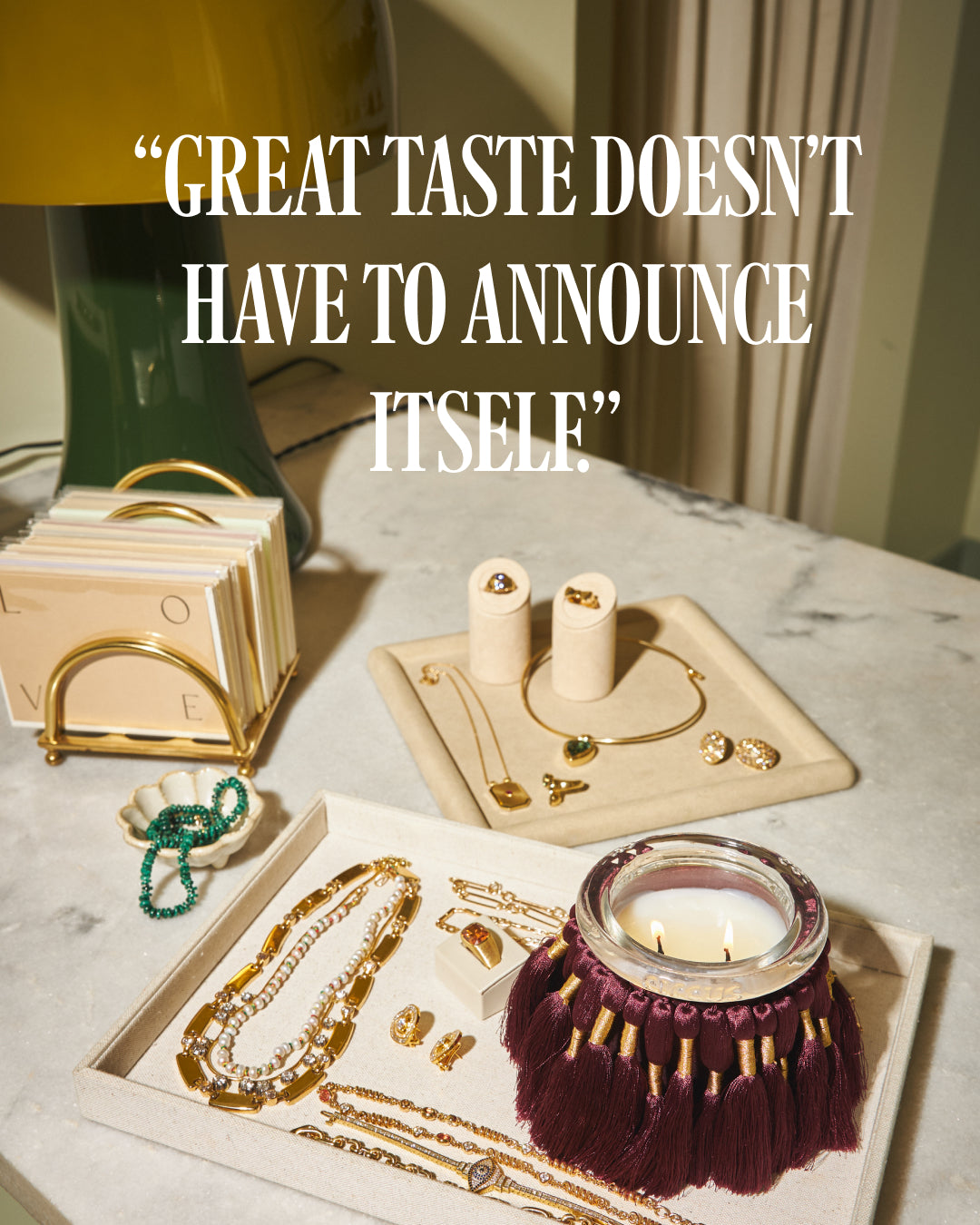 A styled tabletop arrangement of jewelry and home decor with fuzzed by circus candles on a marble surface, with large overlaid text reading, “GREAT TASTE DOESN'T HAVE TO ANNOUNCE ITSELF.” Visible items include gold-tone necklaces, bracelets, earrings, candle holders, a green tassel candle, and a gold card holder.