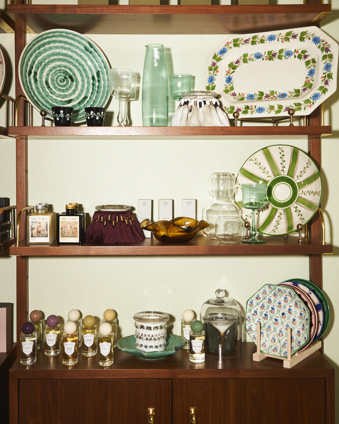 A wooden display cabinet with four shelves holding assorted decorative tableware, glassware, plates, fuzzed circus candle, crystal fetish circus candle and small bottles against a pale green wall.