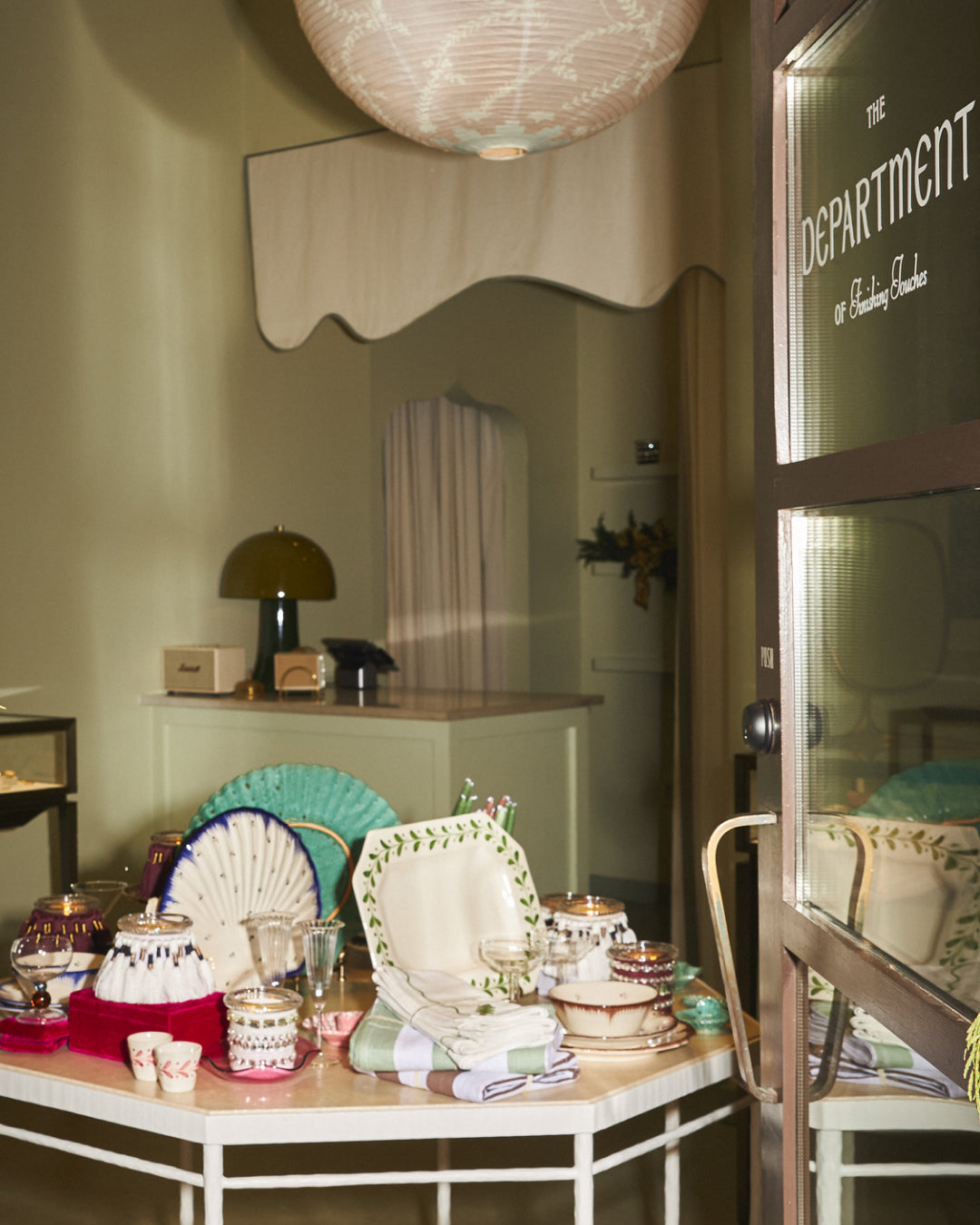 A styled interior scene showing a table set with folded linens, cups, and decorative fans in soft pastel tones, with a mirror, lamp, and a glass door marked 'THE DEPARTMENT' in the background.