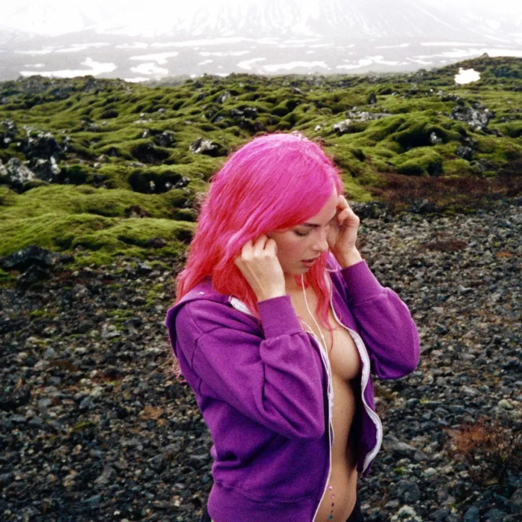 woman with bright pink hair wearing a purple jacket in a mossy landscape